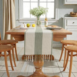 Puede incluir: Una mesa de comedor de madera con un camino de mesa a rayas verdes y blancas. La mesa está decorada con un jarrón de flores blancas, un plato de comida y una botella. Cuatro sillas de madera con asientos de mimbre rodean la mesa.