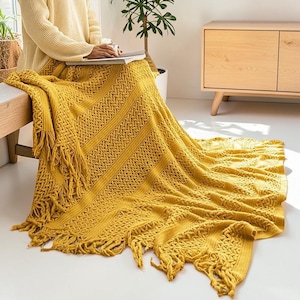 May include: A mustard yellow knitted throw blanket with a textured pattern and fringe detailing. The blanket is draped over a person's lap and extends onto the floor. The throw is made of a soft material and is suitable for home decor.