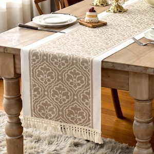 Op de afbeelding: Beige en wit patroon tafelloper met een gefranjerde rand. De loper heeft een herhalend bloemenontwerp en is geplaatst op een houten tafel. De tafel is gedekt met een bord, bestek en een klein dessert.