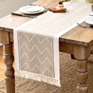 Könnte beinhalten: Beige-weiß gemusterter Tischläufer mit geometrischem Design. Der Läufer hat einen Fransenrand und liegt auf einem Holztisch. Der Tisch ist mit Tellern, Besteck und einem kleinen Dessert gedeckt.