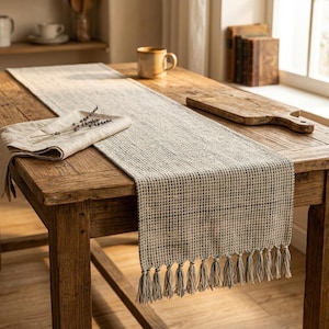 Può includere: Runner da tavolo beige intrecciato con bordo a frange, posto su un tavolo di legno rustico. Un tovagliolo di lino piegato con rametti di lavanda poggia sul runner. Una tazza e un tagliere sono anche sul tavolo.