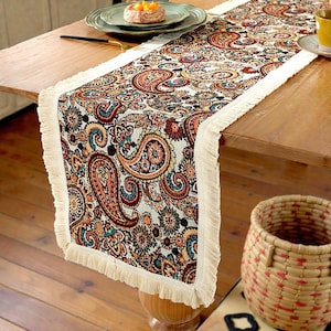 May include: A paisley table runner with a cream fringe. The runner displays a detailed pattern in shades of orange, red, teal, and brown. It is placed on a wooden table, adding elegance to the dining area.