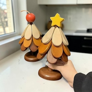 Op de afbeelding: Twee houten kerstboomversieringen. Eén heeft een kers bovenop, de andere een gele ster. De bomen zijn gemaakt van afgestudeerde houten bloembladen in bruin- en beigetinten, gemonteerd op een ronde houten basis.