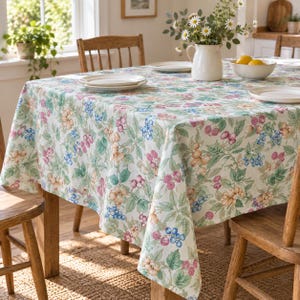 May include: A rectangular tablecloth featuring a botanical print of green leaves, pink raspberries, blue berries, and light orange flowers on a white background. The fabric drapes over a wooden table set with white plates and a white ceramic pitcher.