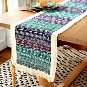 Puede incluir: Un camino de mesa colorido con un patrón geométrico en tonos turquesa, morado y burdeos. El camino de mesa tiene flecos color crema en los bordes. Se coloca sobre una mesa de madera, agregando un toque decorativo al comedor.