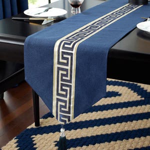Peut inclure: Chemin de table bleu marine avec un motif de clé grecque doré et blanc au centre. Le chemin de table est en tissu texturé et possède un pompon à l'extrémité. Le chemin de table mesure environ 183 cm de long.