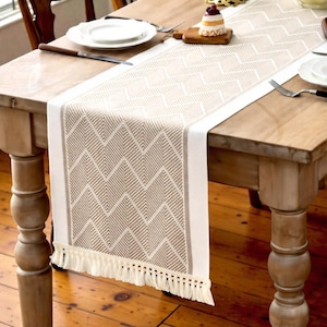 May include: Beige and white table runner with a geometric chevron pattern and fringe trim. The runner is displayed on a wooden table, showcasing its design and texture. The table is set with plates and silverware.
