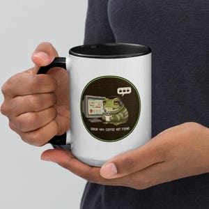 May include: White ceramic mug with a black interior and handle. The mug features a green circular design with a cartoon frog wearing a headset and holding a coffee cup in front of a computer screen. Text on the mug reads "ERROR 404: COFFEE NOT FOUND."