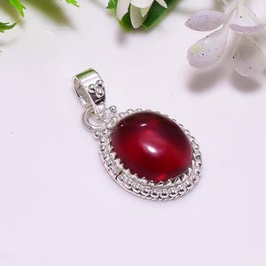 May include: A silver pendant with an oval, deep red gemstone. The gemstone is set in a decorative, beaded silver frame. The pendant has a loop at the top for a chain. The background is white with some green and white floral accents.