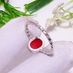 Op de afbeelding: Zilveren ring met een gedraaide band en een ronde, rode edelsteen. De edelsteen is gezet in een zilveren bezel. De ring wordt gepresenteerd op een wit oppervlak met groene bladeren en witte bloemen op de achtergrond.