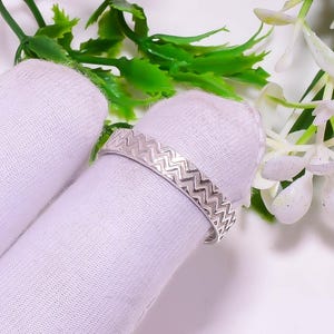 Puede incluir: Un anillo plateado con un patrón en zigzag. El anillo se muestra sobre una tela blanca, con hojas verdes y flores blancas en el fondo. El diseño del anillo es una serie de chevrones conectados.