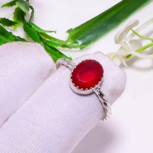 Op de afbeelding: Een zilveren ring met een grote, ovaalvormige, dieprode edelsteen. De edelsteen is gezet in een geschulpte bezel. De band heeft een gedraaid ontwerp. De ring wordt gepresenteerd op een wit oppervlak met groene en witte bloemaccenten.