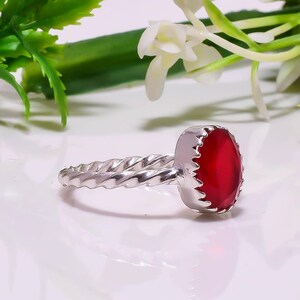 Op de afbeelding: Een zilveren ring met een gedraaide band en een grote, ovale, rode edelsteen. De edelsteen is gezet in een geschulpte bezel. De ring wordt gepresenteerd op een wit oppervlak met groene en witte bloemaccenten.