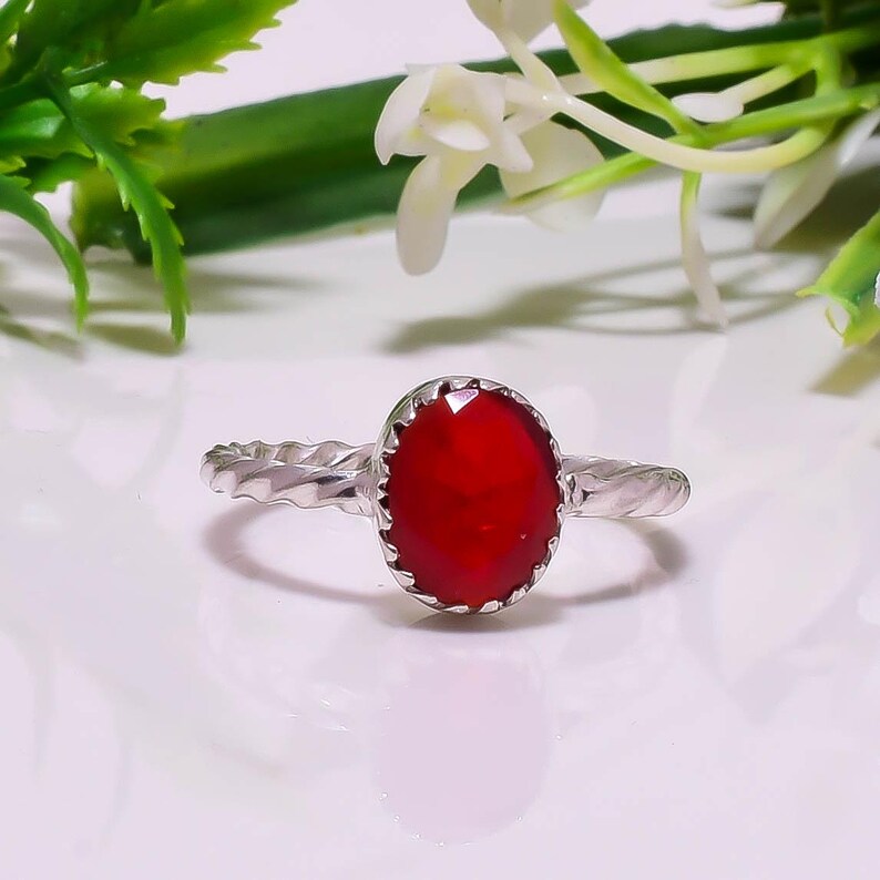 Op de afbeelding: Een zilveren ring met een ovale, dieprode edelsteen. De edelsteen is gezet in een getextureerde bezel en de band heeft een gedraaid ontwerp. De ring wordt getoond op een wit oppervlak, met groene en witte bloemaccenten op de achtergrond.