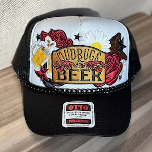 May include: Black and white trucker hat with the text "MUDBUGS and BEER" in gold lettering. The hat features a beer mug, red roses, and a daisy design on the front. The hat has a black brim and mesh back.