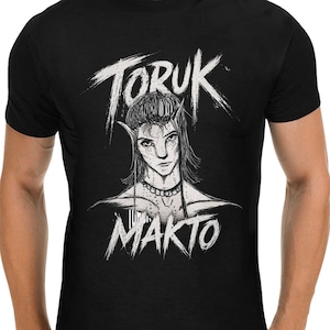 May include: Black t-shirt featuring a white graphic design. The design includes the text "TORUK" above an illustration of a character with pointed ears and the text "MAKTO" below. The graphic has a hand-drawn style.