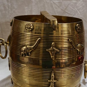 Könnte beinhalten: Ein goldener Messing-Eimer mit Griff und dekorativen Tier- und Blumenreliefs. Der Eimer hat eine zylindrische Form mit horizontalen Rippen und Ringgriffen an den Seiten. Die Schnitzereien umfassen Elefanten und Blumen.