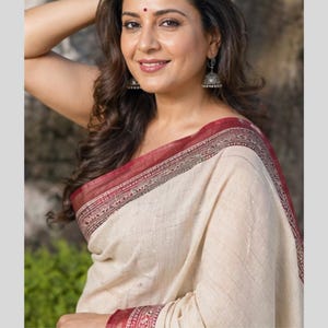 Puede incluir: Una mujer con un sari color crema con borde granate. El sari tiene un sutil estampado de rayas. La mujer sonríe y lleva pendientes plateados. Tiene un bindi rojo en la frente y su cabello oscuro está peinado con ondas.