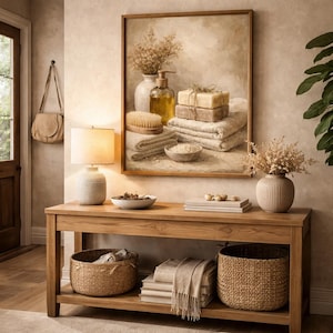 Pu&ograve; includere: Una consolle in legno con due cesti intrecciati, una lampada e un quadro incorniciato. Il dipinto raffigura una natura morta di prodotti per il bagno, tra cui sapone, asciugamani e una spazzola. La scena &egrave; immersa in toni caldi e neutri.