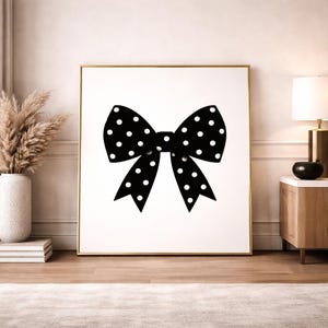 Op de afbeelding: Een ingelijste kunstdruk met een grote zwarte strik met witte stippen. De strik is gecentreerd op een witte achtergrond, goudkleurig omlijst en tentoongesteld in een kamer met neutrale tinten. Het ontwerp van de strik is eenvoudig maar opvallend.