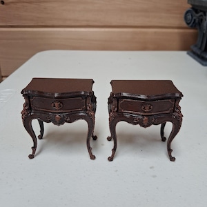May include: Two miniature brown nightstands with ornate detailing. Each nightstand has a drawer and curved legs. The tops are a darker shade of brown than the rest of the piece. The nightstands are placed side by side.