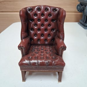 May include: A small, reddish-brown wingback chair with a button-tufted design. The miniature chair has rolled arms and a wooden frame. Ideal for a doll's house or miniature display, this piece is a classic design.
