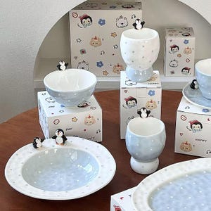 Puede incluir: Un conjunto de vajilla de cerámica en azul claro y blanco con pequeñas figuras de pingüinos en los bordes. El set incluye cuencos, tazas y platos con lunares blancos, acompañados de cajas a juego decoradas con ilustraciones de dibujos animados.