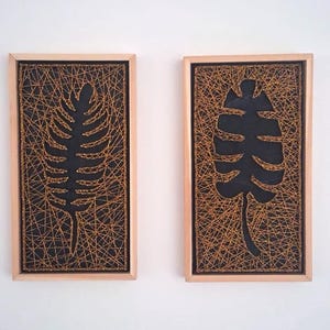 Peut inclure: Deux œuvres d'art encadrées avec des motifs de feuilles. Chacune présente un fond noir avec une silhouette de feuille, recouverte de fil doré. Les cadres sont en bois clair, créant un contraste. Idéal pour la décoration murale.