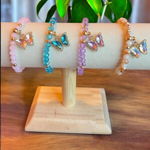 Könnte beinhalten: Vier Armbänder mit Perlen in Rosa, Blau, Lila und Pfirsich, jeweils mit einem goldenen Schmetterlingsanhänger. Die Armbänder werden auf einem Schmuckständer aus Holz präsentiert und zeigen die verschiedenen Farben und Designs.