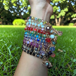 Könnte beinhalten: Mehrere farbenfrohe Armbänder mit Perlen, goldenen Akzenten und hängenden Anhängern. Die Armbänder weisen eine Vielzahl von Perlen in Blau-, Grün-, Rot-, Lila- und Klartönen auf, einige mit Evil-Eye-Designs.