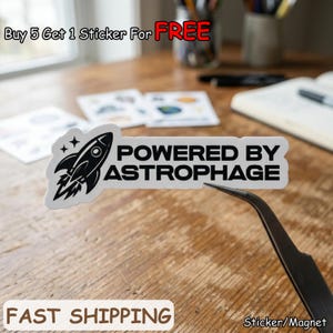 May include: A white sticker with a black rocket ship graphic and the text "POWERED BY ASTROPHAGE." The sticker is being held by tweezers. Additional text includes "Buy 5 Get 1 Sticker For FREE" and "FAST SHIPPING."