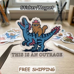 May include: A sticker or magnet featuring a cartoon illustration of a blue dinosaur with pink accents, carrying a brown cat wearing sunglasses and a crown. The text "THIS IS AN OUTRAGE" is below the image.