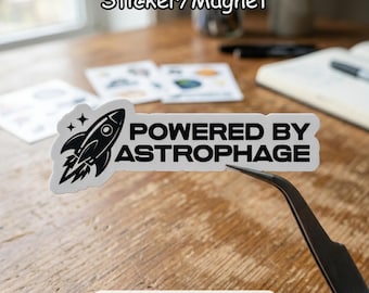 May include: A white sticker/magnet with a black rocket ship graphic and the text "POWERED BY ASTROPHAGE". The sticker is being held by tweezers. Other stickers are in the background.