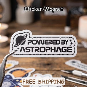 May include: A white die-cut sticker or magnet featuring a black graphic of a planet with rings and the text "POWERED BY ASTROPHAGE" in a bold, modern font. The design includes small four-pointed star accents on the left and right sides.