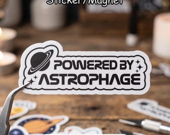May include: A white die-cut sticker or magnet featuring a black graphic of a planet with rings and the text "POWERED BY ASTROPHAGE" in a bold, modern font. The design includes small four-pointed star accents on the left and right sides.