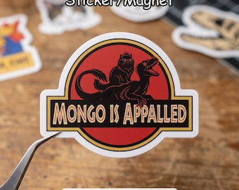 May include: A sticker or magnet featuring a red circle with a black cat and dinosaur illustration. The text "MONGO IS APPALLED" is in a yellow and black rectangular banner. The sticker has a white border and is being held by tweezers.