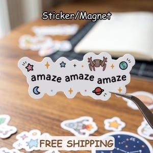 May include: A white die-cut sticker or magnet featuring the repeated black text "amaze amaze amaze" surrounded by colorful space-themed illustrations including a pink star, a blue star, a brown robot, a planet, a crescent moon, and a small Earth.