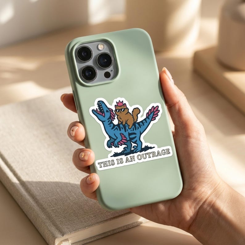 May include: A sticker on a mint green phone case features a cartoon cat wearing a crown riding a blue dinosaur. The dinosaur has pink accents. The sticker has the text "THIS IS AN OUTRAGE" in a white rectangle.