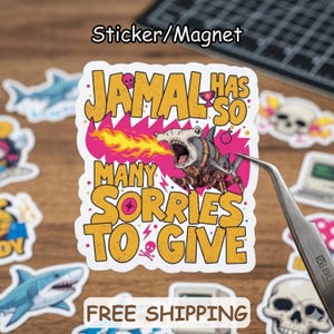 May include: A sticker/magnet with the text "JAMAL HAS SO MANY SORRIES TO GIVE" and a cartoon shark breathing fire. The design is in yellow, pink, and gray. Other stickers with sharks and skulls are visible.