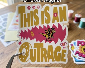 May include: A sticker or magnet with the text "THIS IS AN OUTRAGE" in gold lettering. The design includes a dinosaur riding another dinosaur, a pineapple, and other graphic elements. The sticker is held by tweezers.