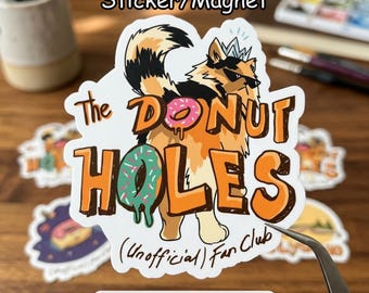 May include: A sticker or magnet featuring a raccoon character with sunglasses and a crown, holding a donut. The text reads "The DONUT HOLES (Unofficial) Fan Club". The design includes a pink and a green donut.