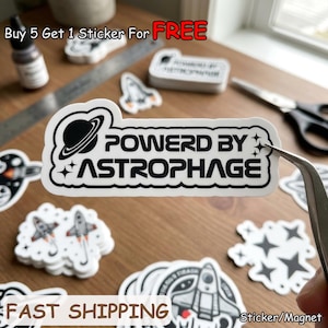 May include: A collection of white stickers with black graphics and text, including a sticker that says "POWERD BY ASTROPHAGE" with a planet and star design. Other stickers feature rockets and stars. The words "FAST SHIPPING" are also visible.