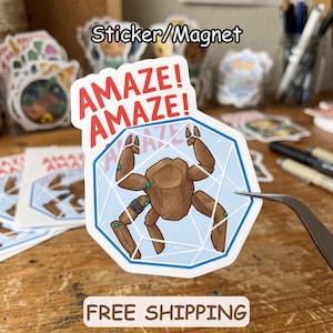 May include: A die-cut sticker or magnet featuring a brown, multi-legged creature inside a light blue geometric shape. The text "AMAZE! AMAZE!" is printed in red above the illustration. The design includes thin white lines forming a polyhedral pattern over the light blue background, with the creature depicted in a segmented, robotic style.