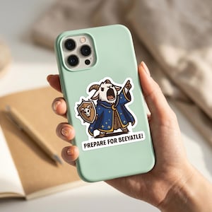 May include: A mint green phone case with a sticker depicting a cartoon goat in a wizard's robe, holding a shield. The goat is shouting and pointing. The sticker text reads, "PREPARE FOR BEEYATLE!"
