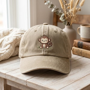 May include: A tan baseball cap featuring an embroidered graphic of a small brown monkey sitting on a patch of green grass. The cap has a curved brim, a button on the top, and stitched eyelets for ventilation, showcasing a classic six-panel design.