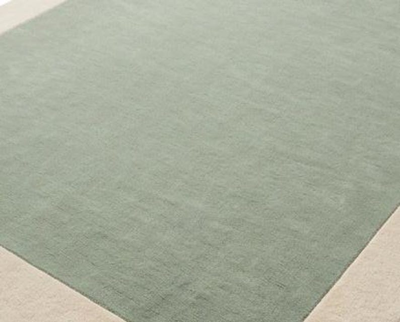 K&ouml;nnte beinhalten: Ein rechteckiger Teppich mit einer gro&szlig;en salbeigr&uuml;nen Mittelfl&auml;che, die von einem einfarbigen cremefarbenen Rand umgeben ist. Der Teppich hat eine kurze, dichte Florstruktur, die auf der gesamten Oberfl&auml;che gleichm&auml;&szlig;ig erscheint und ein schlichtes Design bildet.