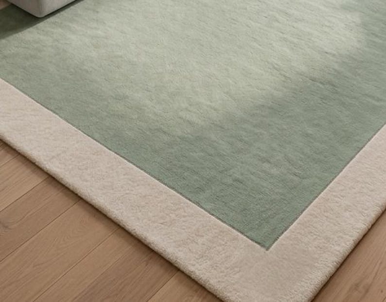 K&ouml;nnte beinhalten: Ein rechteckiger Teppich mit einem gro&szlig;en salbeigr&uuml;nen Mittelfeld, das von einem breiten, cremefarbenen Rand umgeben ist. Der Teppich hat eine weiche, strukturierte Floroberfl&auml;che und liegt flach auf einem hellen Holzboden.
