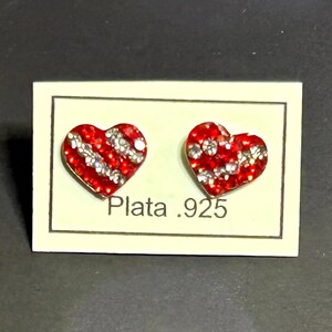 Puede incluir: Un par de pendientes de botón en forma de corazón. Los pendientes son rojos con detalles plateados y están colocados sobre una tarjeta crema. La tarjeta tiene el texto "Plata .925" impreso.