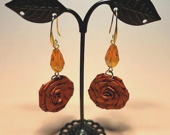May include: A pair of dangling earrings featuring a brown rose-shaped charm made of woven material, suspended from a gold-toned hook with a faceted amber-colored teardrop bead positioned directly above the rose charm.