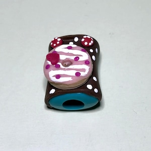 May include: A whimsical pencil sharpener featuring a donut design. The donut is pink with white icing and pink sprinkles, and a heart-shaped decoration. The sharpener's body is brown with white polka dots and a teal base.
